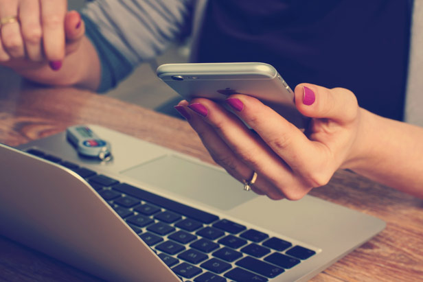 Person mit pinken Fingernägeln hält ein iPhone in der Hand und sitzt vor einem MacBook