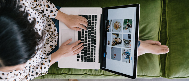 Eine Frau sitzt mit ausgestreckten Beinen auf einem grünen Sofa. Sie hat ein MacBook auf dem Schoß, auf dessen Bildschirm zu erkennen ist, dass die Frau an einer Videokonferenz teilnimmt. Das Bild ist aus der Vogelperspektive aufgenommen